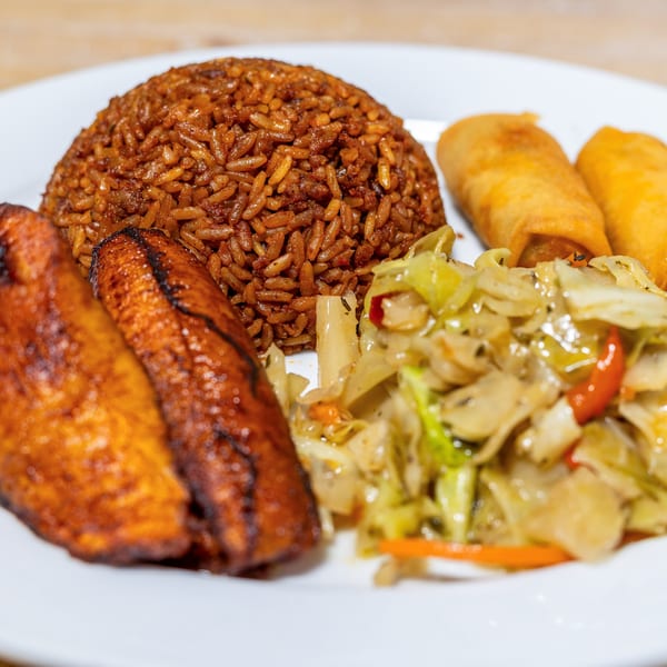 A Caribbean meal with rice, plantains, cabbage, and two spring rolls on a white plate.