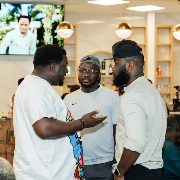 Three men conversing in a brightly lit Caribbean restaurant with a TV screen in the background.